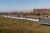 A white train is stopped in a field near a factory building with steam stacks. The train has "Tranvasco" written on the side. Orange flags are set along the train in the field to signal vaccine centers.