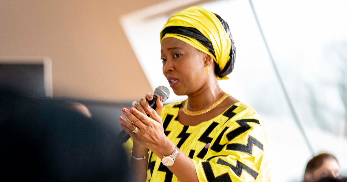 Fatima Maada Bio speaks into a microphone to a group of people. She is wearing a robe and headdress that are both bright yellow with black highlights.