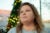 A woman glances off camera. A blurred Christmas tree and outdoor plaza are in the background.