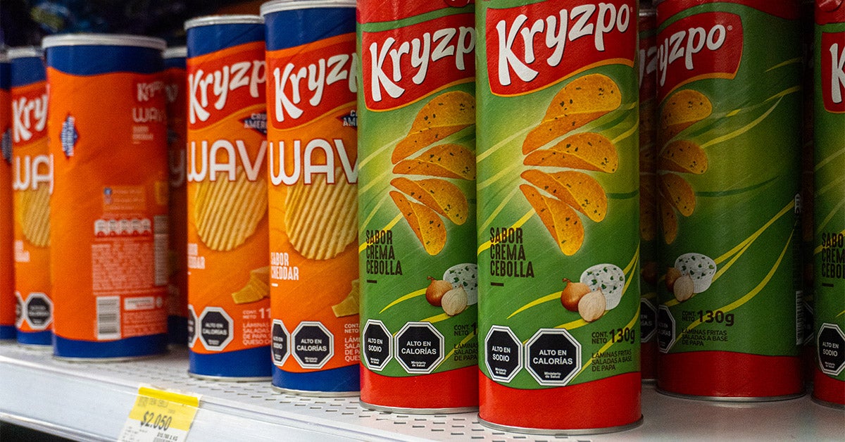 Cans of Kryzpo brand potato chips, labeled in Spanish, line a supermarket shelf. Black labels on each can warn they are "high in sodium" and "high in calories."