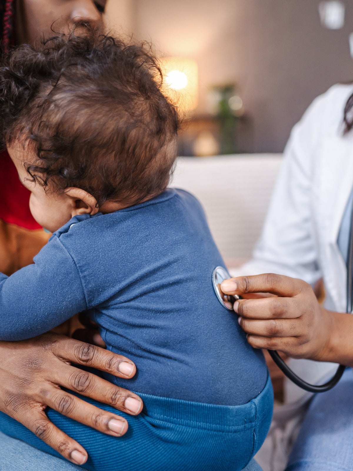 A black female healthcare worker visits a mother and son at their home. The healthcore worker checks his lungs with a stethoscope.