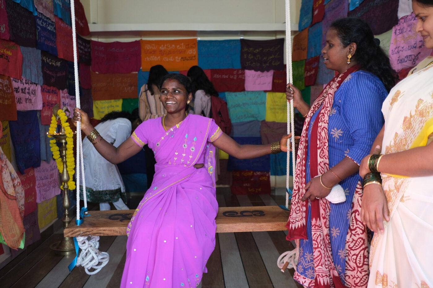 An Indian woman in a bright magenta sari smiles as she sits on a wooden swing in a small gallery space. On the walls are pieces of colorful square fabric with hand-stitched messages.