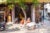 Multiple women sit in a store stall on an indian business street. In the stall, there is a swing and clay casts of ears on the walls. The stall is next to a bodega with chips and other food. A child plays in the foreground, and a tree grows in the middle of the street.