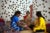 Three Indian women sit on straw mats in a gallery room with colorful pillows. On the walls are clay-cast ears.