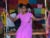 An Indian woman in a bright magenta sari smiles as she sits on a wooden swing in a small gallery space. On the walls are pieces of colorful square fabric with hand-stitched messages.