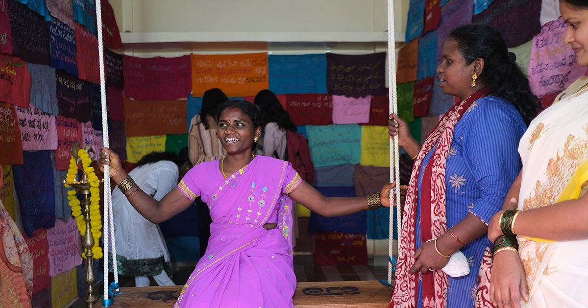 An Indian woman in a bright magenta sari smiles as she sits on a wooden swing in a small gallery space. On the walls are pieces of colorful square fabric with hand-stitched messages.