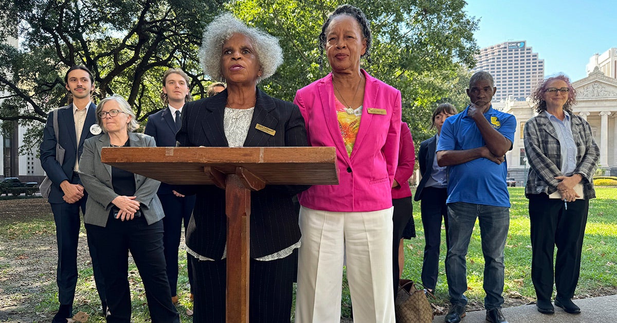 Gail LeBoeuf and Barbara Washington stand at a podium. New Orleans buildings and a large tree are visible in the background.