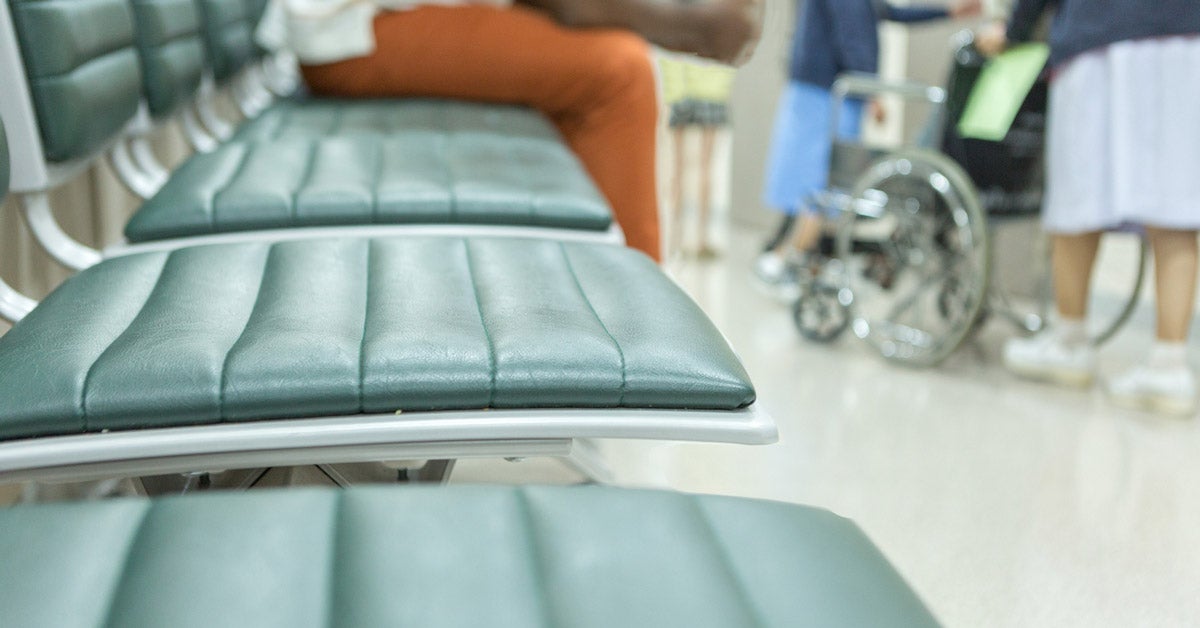 A man sits in a row of waiting room seats while nurses and wheel chairs move through a busy hallway at a hospital.