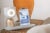EllieQ, a desktop-sized device with a gold base and white oval shape on top, glows with a ring of white light. A tablet and camera are attached to the base on its right. EllieQ sits on a wicker side table with an orange vase in the background.