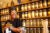 Angela Mallet smiles widely off-camera while standing behind the counter at her general shop in Milwaukee, Wisconsin. Shelves with glass jars with yellow labels holding goods are behind her. She wears a black tshirt, large earrings, bright yellow glasses and a headwrap.