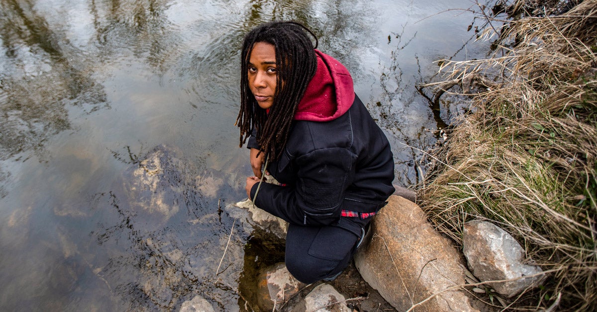 Marcel Simmons squats on some rocks near the edge of the Flint River. He stares somberly up at the camera.