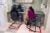 Two female patients sit in front of a plexiglass window at a medical clinic. They speak to a female worker wearing a grey cardigan and mask.