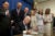 An elected government official signs a bill on a large wooden table. Colleagues and constituents stand behind him, looking-in.