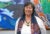 Jane Garcia smiles at the camera and stands in front of a colorful mural. She wears a long sleeved white blouse, navy and orange patterned scarf, teal stone necklace and wears her hair short.
