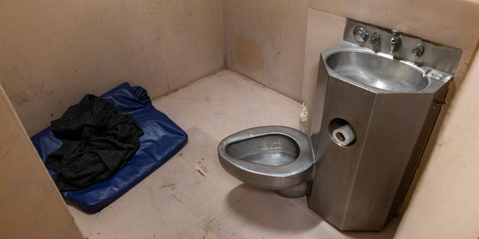 A padded jail cell with filthy, yellowing walls and a dirty floor has a metal sink and toilet attached to the wall. In the corner is a small blue sleeping bag and black blanket.