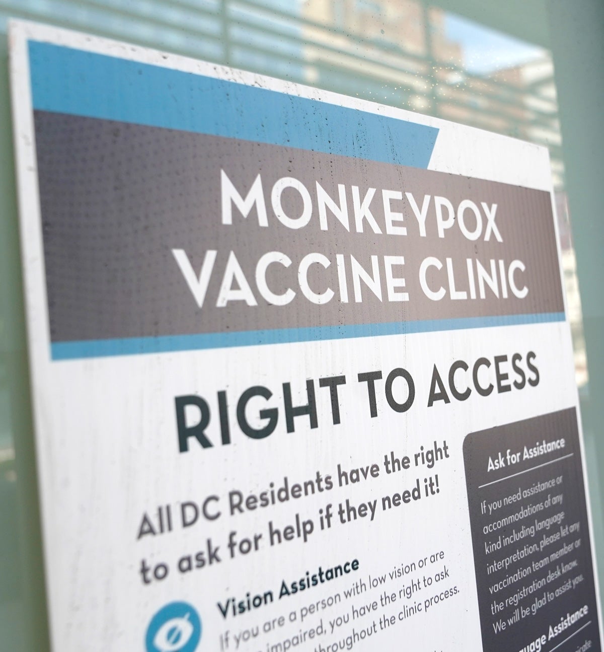A blue, white and gray sign reads “Monkeypox Vaccine Clinic Right To Access”. It it taped to a storefront window. A “DC Health” logo is affixed to an entrance door in the lower right-hand corner of the image.