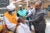 Heitham Mohammed Ibrahim Awadalla, Sudan’s minister of health, administers an oral vaccine to an adult man at a vaccine event. A healthcare worker puts his hands ont he patient’s shoulders. Two other people look on and smile, taking photos.