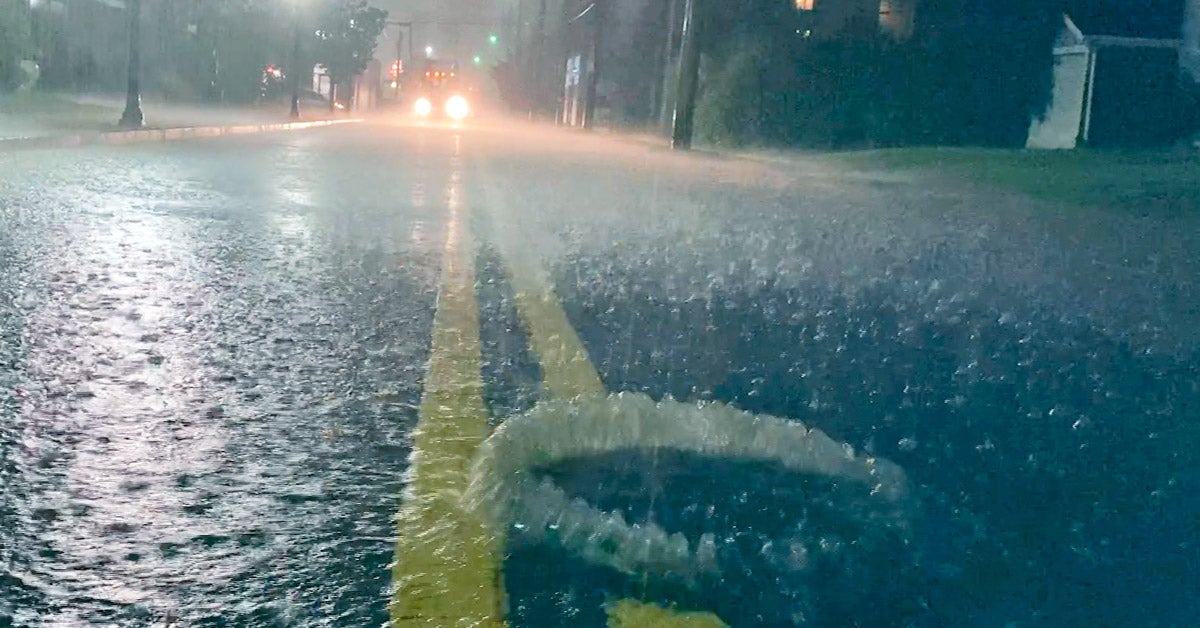 A flooded neighborhood street with a utility hole overflowing with bubbles. Car with headlights approaches.