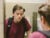 A depressed-looking, white teenage boy wearing a maroon shirt and black backpack looks at himself in a school bathroom mirror.