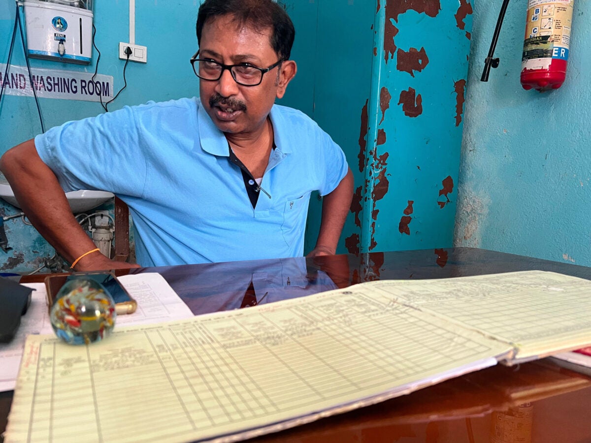 A male indian health worker wearing a blue polo and glasses sits at a desk at a health city with a yellow-paged ledger. The room is also blue, has chipping paint and a hand washing station.