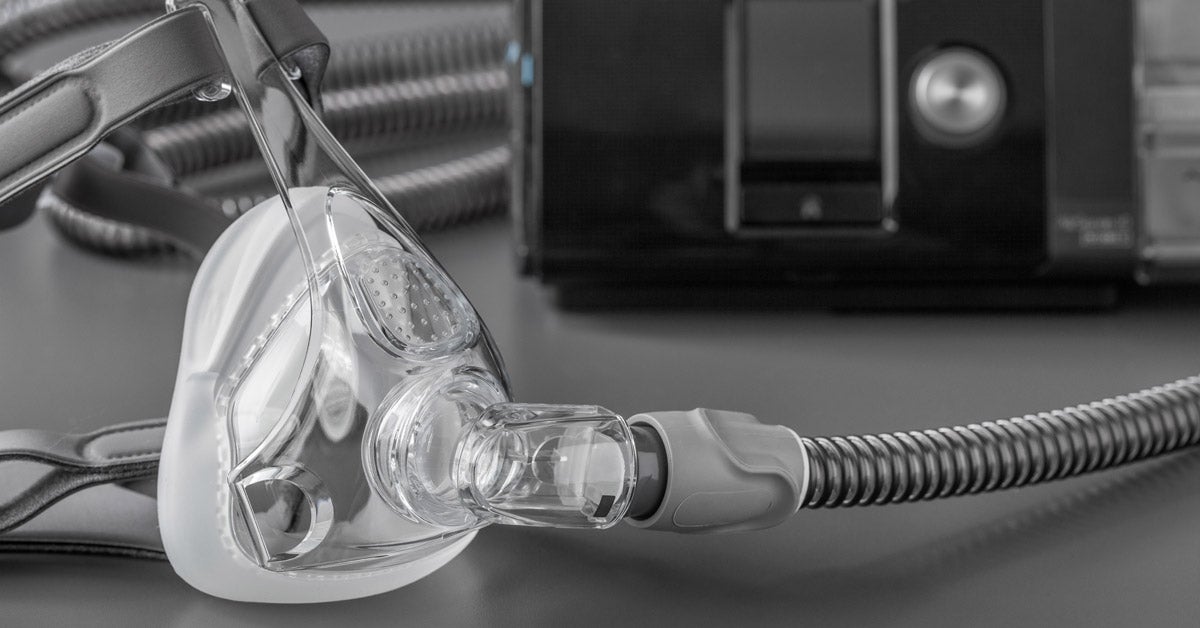 Clear sleep apnea CPAP mask with grey straps and tubes on a grey table. The black CPAP machine is in the background.