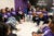 A group of diverse female teens sit in a circle wearing purple t-shirts that read “WOW”. They listen to a middle-aged female in a seat on the right who speaks with her arms outstretched.
