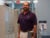 Aaron Dudley wears a purple polo and khakis and stands in a hallway of a medical facility, looking at the camera. He has a stethoscope and key card around his neck.
