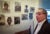 Jack Geiger stands in a hallway of framed black and white historical photographs of black patients. He looks off camera left.