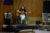 A singer performs with a guitar on a wooden stage to a small, seated crowd.