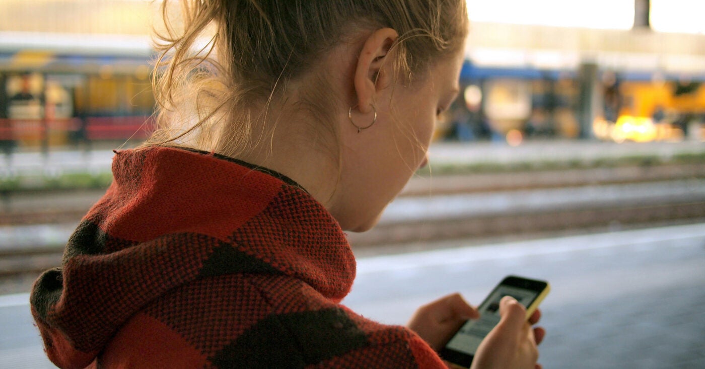 A female teen in a red plaid hoodie interacts with her phone screen at a train station.