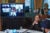 Vice President Harris sits at a table , looking at a large tv screen on her left. Seven people are indivudally shown in a grid for the group zoom call on the screen.