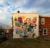 Mural entitled "A Collection of Creative Decisions" Painted on the side of brick house, a white wall is filled with 3D sqaures and shapes, protruding out. They are bright colros and show science and figures: a guitar, a sun, a bird, a woman with an umbrella, a christmas tree, a dog, and others.