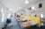 The Butaro District Hospital in Rwanda: a short yellow wall divides a large room with evenly spaced hosptial beds on each side. Mosquito nets hang above each bed. White lights and many windows make the space bright. Patients sit on beds or in wheelchairs along the wall.