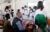 Paul Farmer sits with his back to the camera wearing a blue button down and black vest. He is talking to a group of 10+ African medical professionals wearing lab coats and facemasks. Women wearing traditional African clothing sit on either side of Paul at Butaro District Hospital in Kigali, Rwanda.