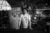 Black and white photo: A twenty-something man stands in his garage, resting his hands on a pooltable. His garage is decorated with stickers, family photos, and native american artifacts. He wears a distressed t-shirt, necklace with an arrowhead and stares directly into the camera.