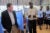 Archival photo, 2012: A tall African man wearing a dress shirt and pants speaks to a white man wearing a dress shirt, pants and blazer in an African clinic. Two clinic workers stand behind them.