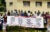 Archival photo, 2003: A group of Ugandan protestors stand in front of a yellow building and hold a large banner that reads "Dear President Bush: Generic drugs are the reason HIV/AIDS have got cheaper. But you say you wont buy generics with you HIV/AIDS money. You will only buy drugs from big pharmaceutical companies. Do you want to save lives or make profits for your industry friend? Ugandan coalition for access to essential medicines.