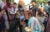 An older woman drinks from a blue plastic cup and holds a plate of food in an open street, surrounded by others who are also drinking.