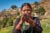A eldery Nepalese woman breathes into an inhaler outside. She wears gold hoop earings and bangles.
