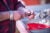 Person wearing a red plaid shirt and silver bracelets holds a knife and shucks an unidentifed shellfish. A untility sink full of shells in on one side and with a red bucket in the other is on the right side of the image.