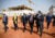 Four governmental officals wearing suits and face masks walk on orange dirt, conversing. A building under construction with a crane and white tacking is behind them. Other government officials and men in uniform walk behind.