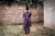 A woman stands with her back to the camera near a brick wall. A freshly dug grave is at her feet.